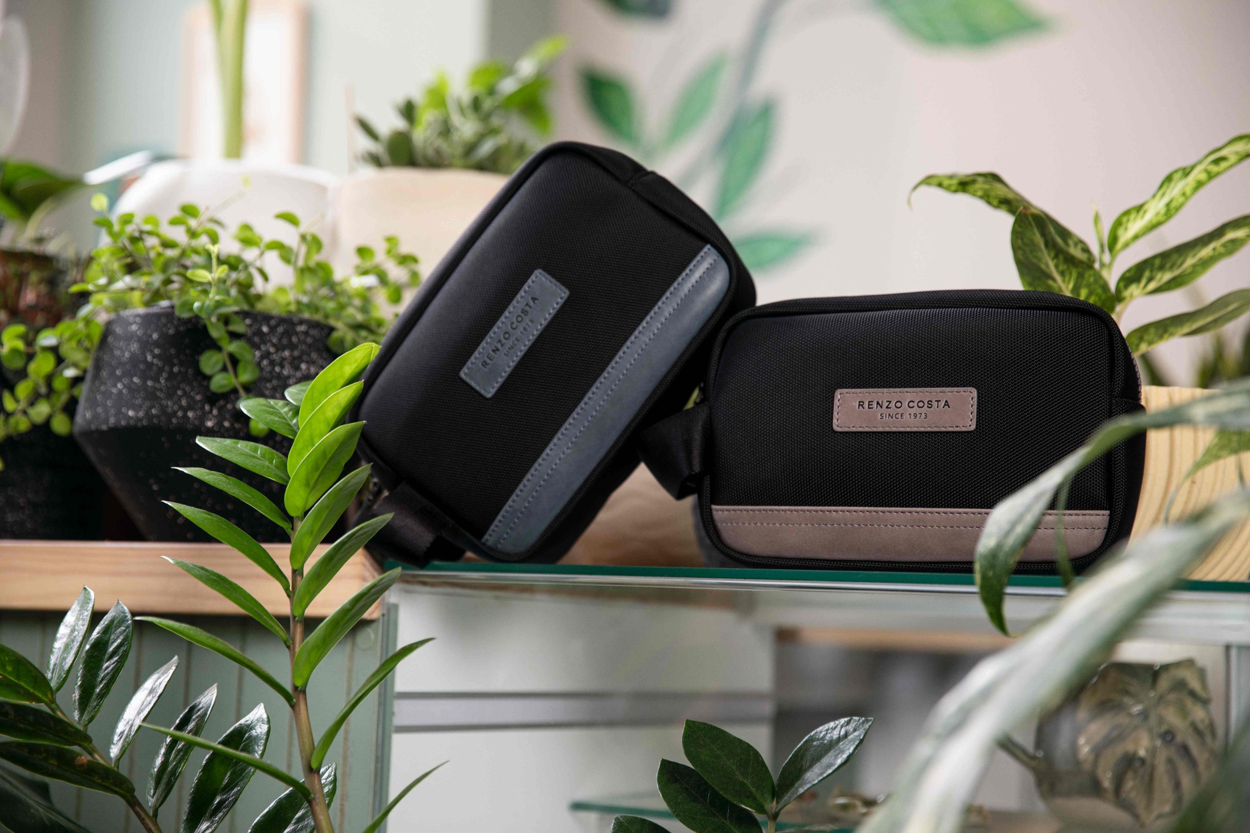 Neceser de nylon negro Renzo Costa Urban Grid con detalles en gris, presentado entre plantas en una composición de producto.