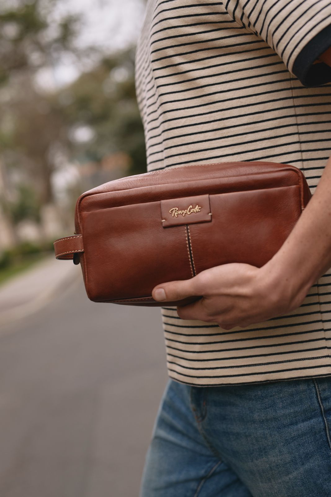 Neceser marrón Renzo Costa para hombre, sostenido en la mano y combinado con un look casual .